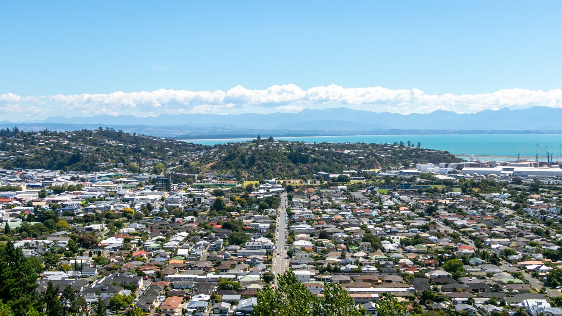 High-angle view of a coastal city with a central road, blue ocean, and mountains.